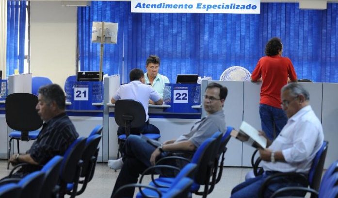 #pracegover: foto mostra agência do INSS, com três pessoas sentadas aguardando para serem atendidas em primeiro plano. Atrás um homem em pé no guichê e outro sentado sendo atendido por profissional do INSS